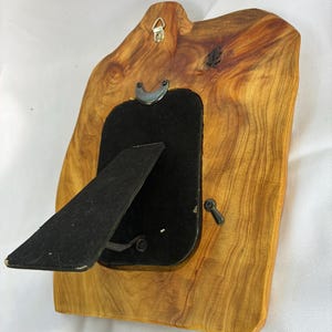 May include: A natural edge wooden picture frame with a light brown grain pattern. The back features a black velvet backing, a black easel stand, a metal hanging hook at the top, and small rotating metal tabs to secure the frame contents in place.