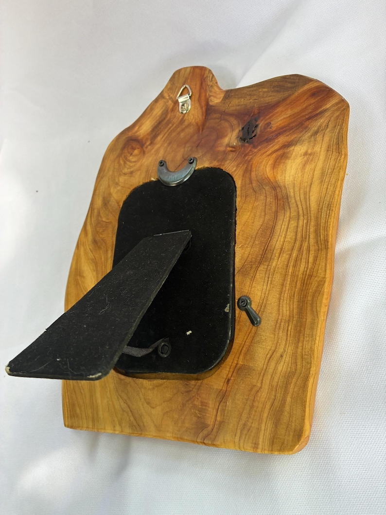 May include: A natural edge wooden picture frame with a light brown grain pattern. The back features a black velvet backing, a black easel stand, a metal hanging hook at the top, and small rotating metal tabs to secure the frame contents in place.