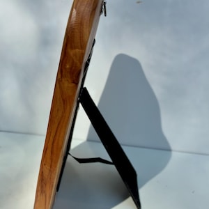 May include: A side view of a wooden picture frame with a smooth, polished finish showing natural wood grain patterns. The frame is supported by a black easel-style stand attached to the back, featuring a metal hanging loop at the top edge.