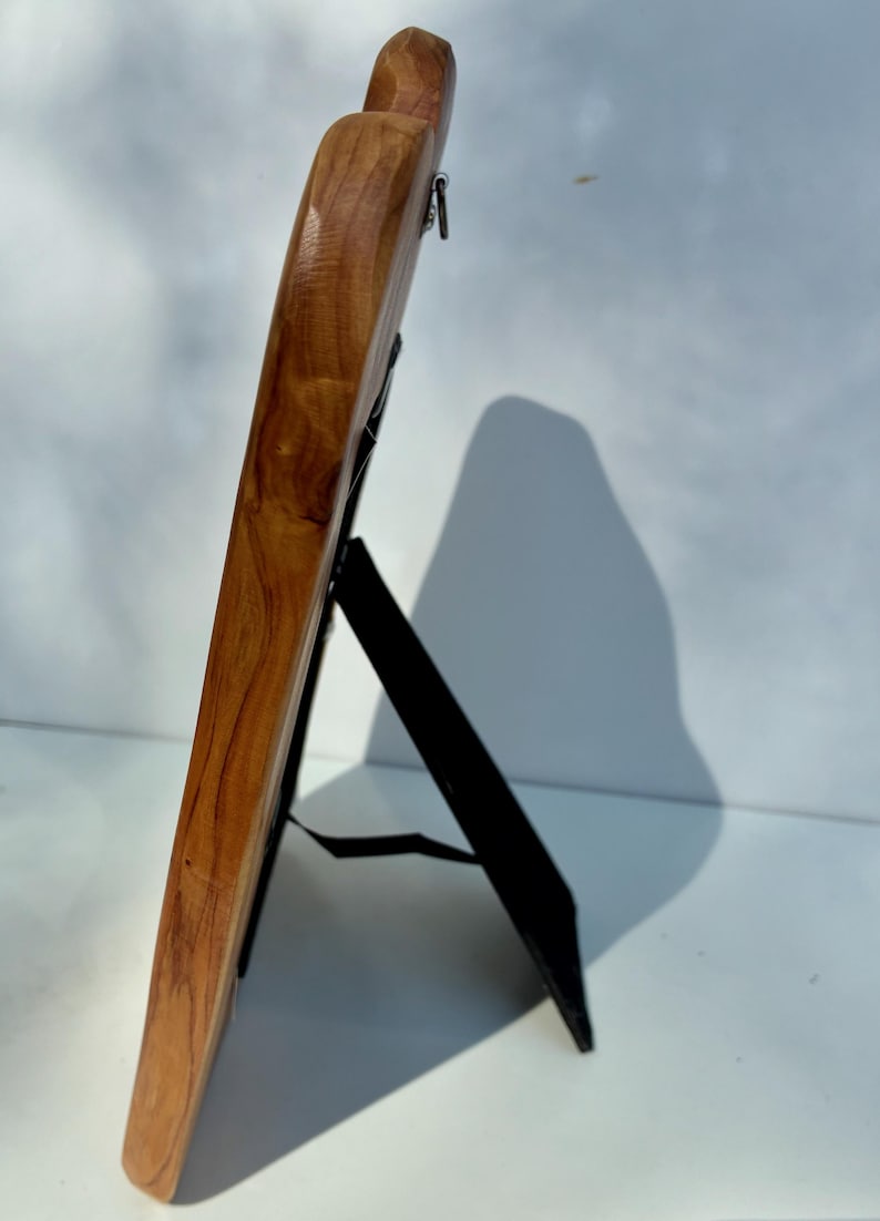 May include: A side view of a wooden picture frame with a smooth, polished finish showing natural wood grain patterns. The frame is supported by a black easel-style stand attached to the back, featuring a metal hanging loop at the top edge.