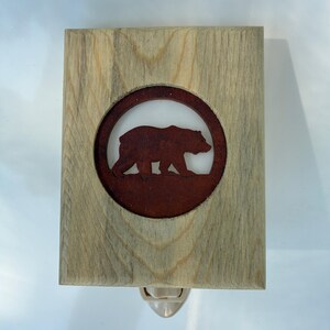 May include: Wooden night light featuring a brown bear silhouette within a circular frame. The bear is set against a white background, framed by a rectangular wooden border. The night light has a standard plug-in base.