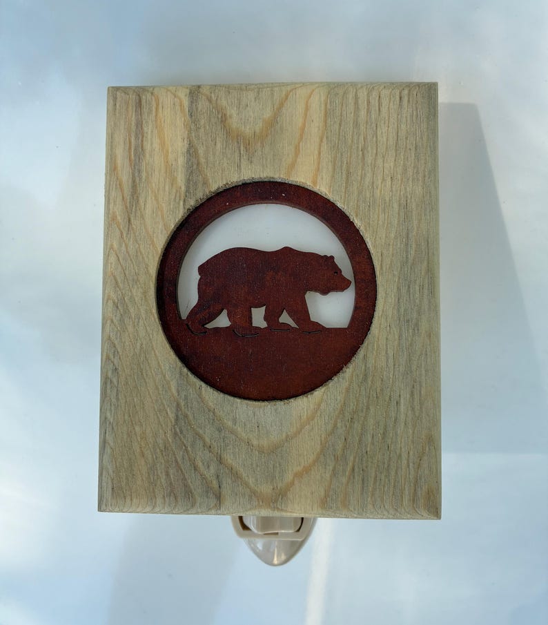 May include: Wooden night light featuring a brown bear silhouette within a circular frame. The bear is set against a white background, framed by a rectangular wooden border. The night light has a standard plug-in base.