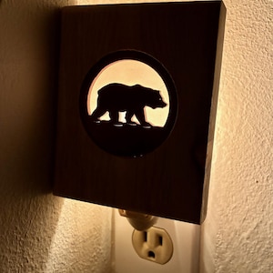 May include: A wooden night light with a cutout of a bear silhouette inside a circle. The night light is plugged into a white wall outlet. The bear is walking to the right, and the light shines through the cutout.