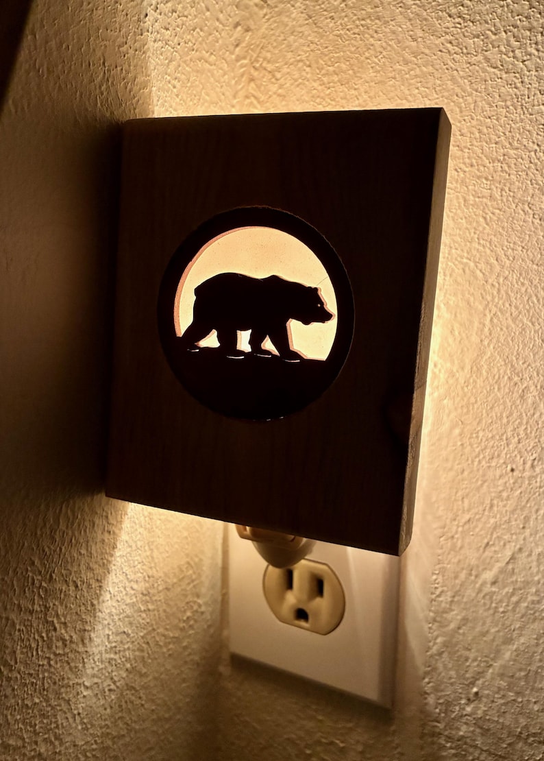 May include: A wooden night light with a cutout of a bear silhouette inside a circle. The night light is plugged into a white wall outlet. The bear is walking to the right, and the light shines through the cutout.