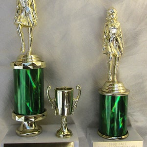 May include: Three trophies with gold figures atop green and gold columns. The largest two have a female figure. The smallest is a gold cup with handles. The bases are white with gold nameplates. One trophy reads "1992 Fall Tavelli Terminators R.J. Moser."