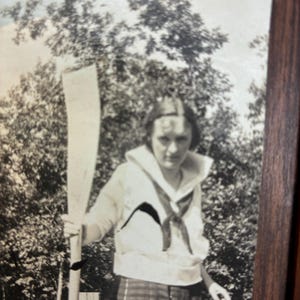 May include: A black and white vintage photograph featuring a person wearing a sailor-style top with a large collar and a dark neckerchief, paired with a plaid skirt. The person is holding a long wooden oar upright in an outdoor setting with trees.