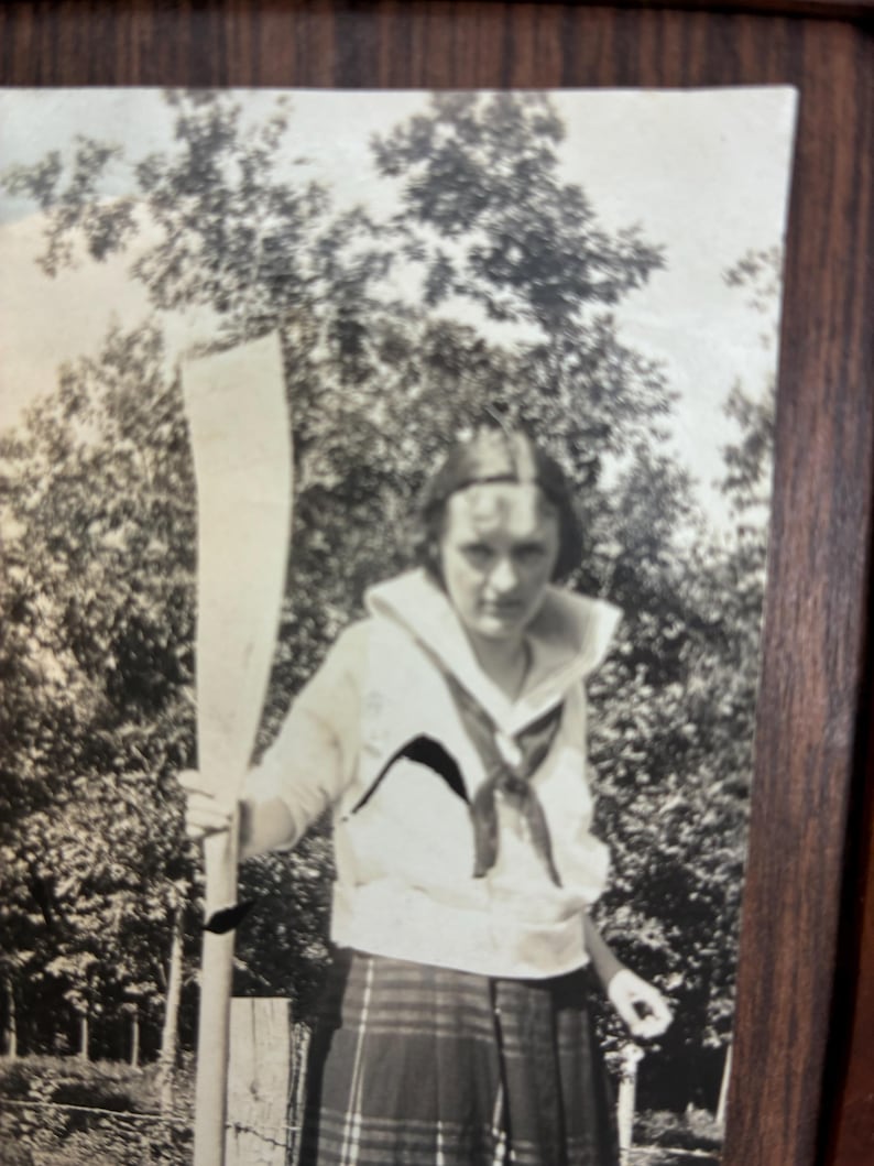 May include: A black and white vintage photograph featuring a person wearing a sailor-style top with a large collar and a dark neckerchief, paired with a plaid skirt. The person is holding a long wooden oar upright in an outdoor setting with trees.