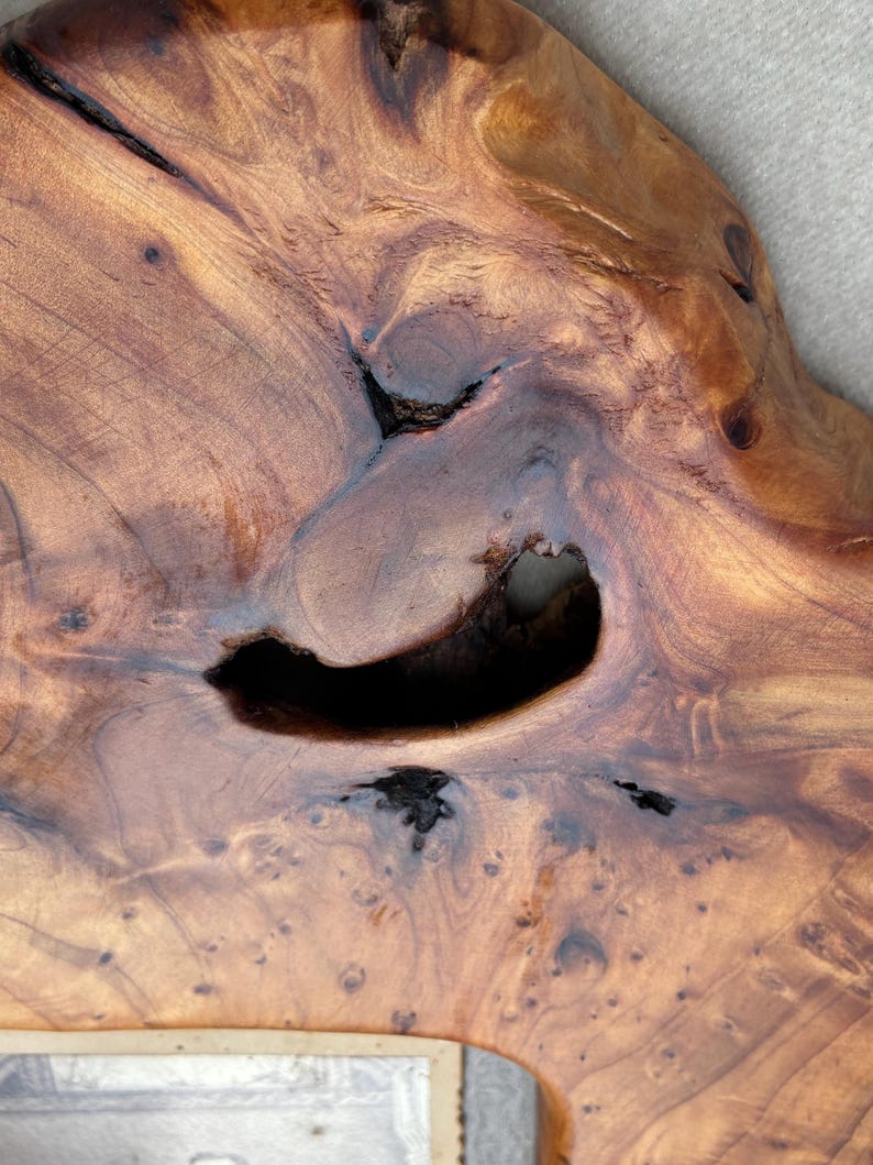 May include: A polished wooden slab featuring a natural live edge and a prominent central void. The surface displays a rich amber and brown grain pattern with dark knots, swirling wood textures, and organic irregularities throughout the piece.