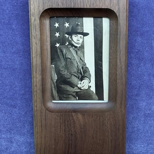 May include: A rectangular dark brown wooden frame with rounded inner corners containing a black and white photograph of a soldier in a uniform and hat, posed in front of an American flag with visible stars and vertical stripes in the background.