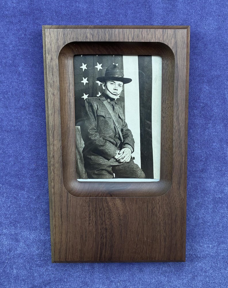 May include: A rectangular dark brown wooden frame with rounded inner corners containing a black and white photograph of a soldier in a uniform and hat, posed in front of an American flag with visible stars and vertical stripes in the background.