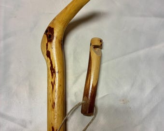 May include: A handcrafted wooden walking stick with a curved handle and a separate whistle. The walking stick is light brown with dark brown accents. The whistle is attached with a natural fiber cord. The wood grain is visible.