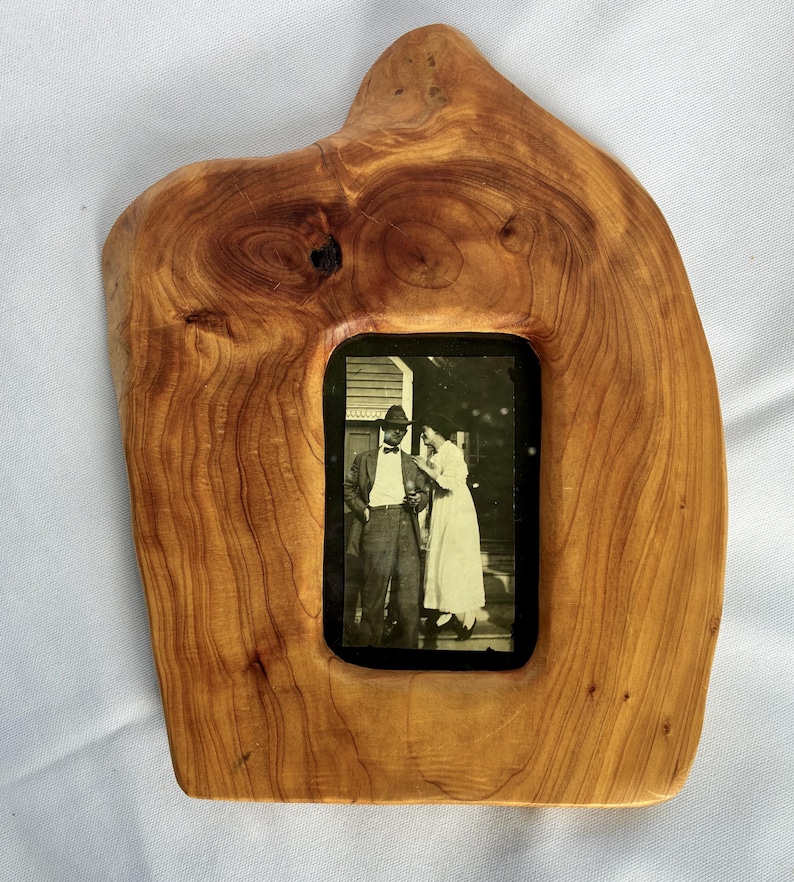 May include: A natural-edged wooden picture frame with a light brown, polished finish showing prominent wood grain patterns and knots. The frame holds a rectangular black and white photograph depicting two people standing outdoors in vintage attire.