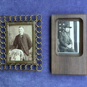 May include: Two rectangular picture frames. The left frame is made of brass with a decorative chain-link border, containing a black and white portrait of a person in a suit. The right frame is made of dark wood, containing a black and white portrait.