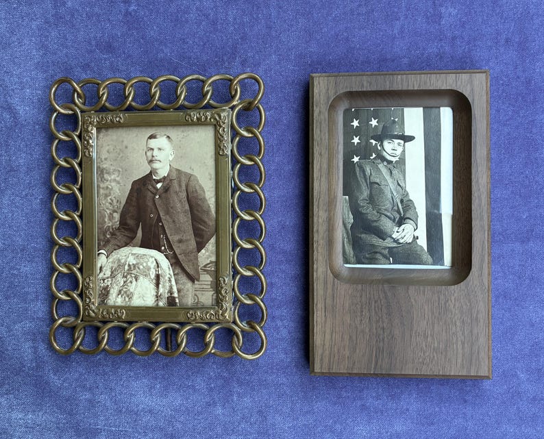 May include: Two rectangular picture frames. The left frame is made of brass with a decorative chain-link border, containing a black and white portrait of a person in a suit. The right frame is made of dark wood, containing a black and white portrait.