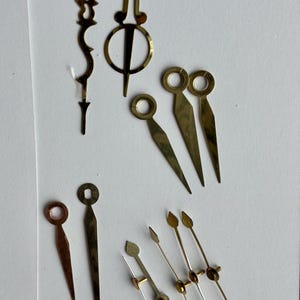 May include: A selection of vintage watch hands in various shapes and sizes. The hands are made of brass and gold-toned metal. Some have circular ends, while others are pointed or decorative. The assortment includes hands for hours, minutes, and seconds.