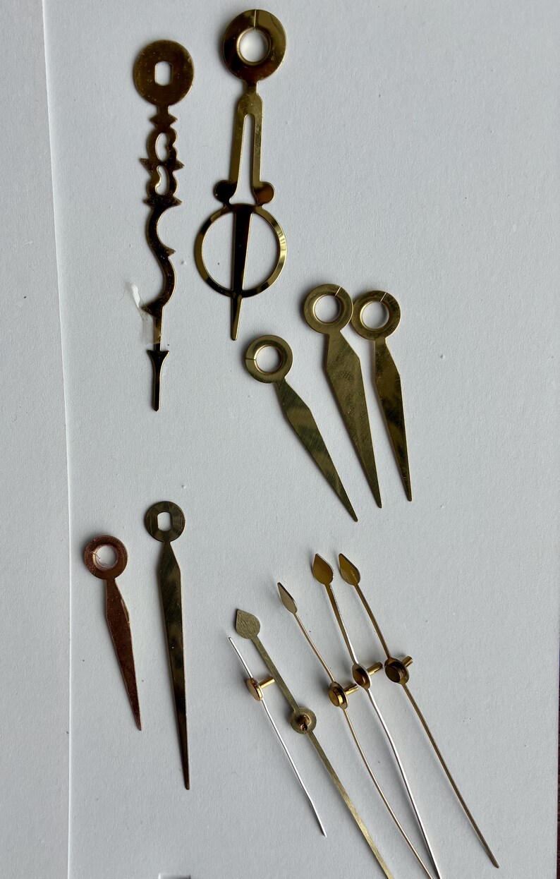 May include: A selection of vintage watch hands in various shapes and sizes. The hands are made of brass and gold-toned metal. Some have circular ends, while others are pointed or decorative. The assortment includes hands for hours, minutes, and seconds.