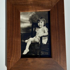 May include: A rectangular wooden picture frame with a dark brown, smooth finish and angled edges. The frame contains a black and white photograph of a young child sitting on a dark chair, wearing a light-colored dress and white shoes.