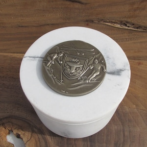 May include: A round, silver-toned metal medallion featuring skiers and a torch emblem, resting on a white marble container. The medallion is engraved with the year "'84". The container is cylindrical with a lid.