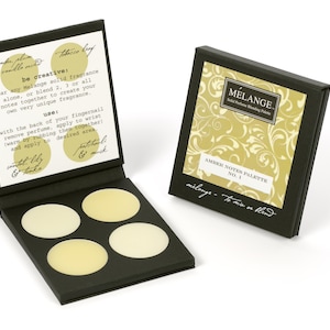 May include: A black box with four round, cream-colored solid perfume samples. The box is open and the lid has instructions for using the perfume. The lid also has the text "be creative: wear any Melange solid fragrance alone, or blend 2, 3 or all notes together to create your own very unique fragrance." The box is labeled "MELANGE Solid Perfume Blending Palette AMBER NOTES PALETTE NO. 1 milange - to mix or blend"