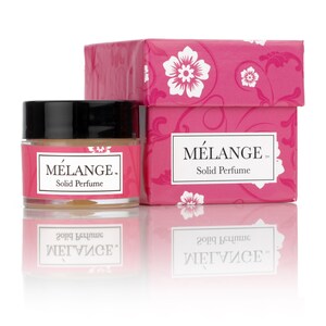 May include: A small, clear jar of solid perfume with a black lid. The jar is labeled "MELANGE. Solid Perfume". The jar is sitting on a white surface in front of a pink box with a floral design. The box is also labeled "MELANGE. Solid Perfume".