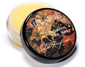 May include: A small round jar of solid perfume with a black lid. The lid has a floral design and the text "MELANGE Perfume Tobacco & Tea Noir Solid Perfume".