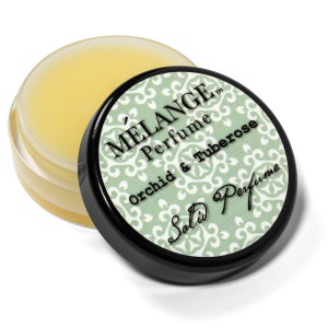 May include: A small, round jar of yellow lip balm with a black lid. The lid has a green and white design with the text "Mélange Perfume Orchid & Tuberose Salti Perfume" in black.