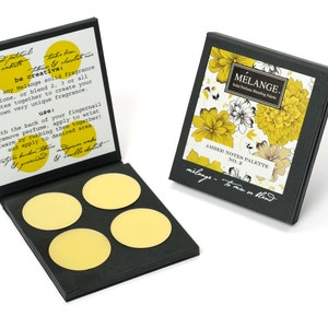 May include: A black box with a floral design on the lid. The box contains four round, yellow solid perfume discs. The text on the box reads "MELANGE. Solid Perfume Blending Palette AMBER NOTES PALETTE NO. 2 melange - to mix or blend".