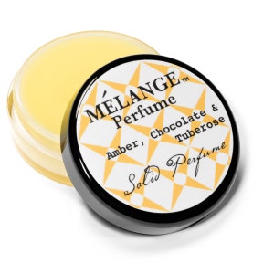 May include: A small, round jar of yellow lip balm with a black lid. The lid has a white label with the text "MÉLANGE™ Perfume Amber, Chocolate & Tuberose Salix Perfume" in black lettering.
