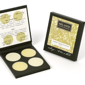 May include: A black box with four round, cream-colored solid perfume samples. The box is open and the lid has instructions for using the perfume. The lid also has the text "be creative: wear any Melange solid fragrance alone, or blend 2, 3 or all notes together to create your own very unique fragrance." The box is labeled "MELANGE Solid Perfume Blending Palette AMBER NOTES PALETTE NO. 1 milange - to mix or blend"