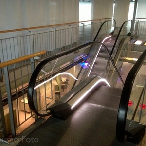 Op de afbeelding: Twee roltrappen met een zilveren leuning en een glazen paneel. De roltrappen bevinden zich in een modern gebouw met een witte muur en een houten leuning.