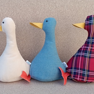 May include: Four plush toy ducks in different patterns. The ducks are sitting in a row on a brown surface. The first duck is black with gold polka dots, the second is white, the third is blue, and the fourth is red with a plaid pattern.