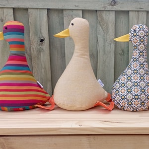 May include: Four plush duck toys lined up on a wooden surface. The ducks are made of different fabrics and have different patterns. One duck is white with a yellow beak, one is striped with red, orange, yellow, and blue, one has a geometric pattern in shades of blue, yellow, and white, and one has a geometric pattern in shades of orange, yellow, and brown.