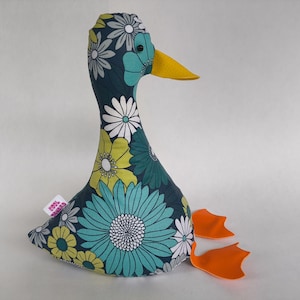 Peut inclure: Un jouet en forme de canard en tissu avec un motif floral bleu foncé orné de fleurs blanches, jaunes et turquoise. Le canard a un bec jaune et des pieds orange. Une petite étiquette est attachée au corps du canard.