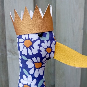 May include: A plush duck toy with a gold crown and a blue and white floral pattern. The duck has a yellow beak and black eyes.