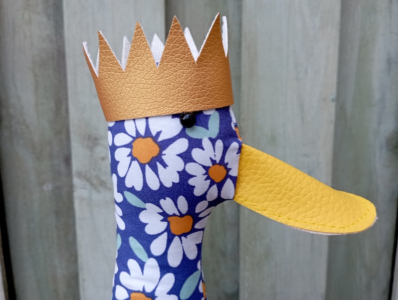 May include: A plush duck toy with a gold crown and a blue and white floral pattern. The duck has a yellow beak and black eyes.