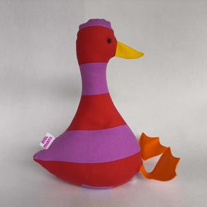 May include: A stuffed duck toy in red and purple stripes with a yellow beak and orange feet. The duck has a small tag that reads "Cool Canvas". The toy is designed with a whimsical and colorful aesthetic.