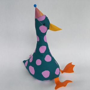 Puede incluir: Un peluche de pato verde azulado con lunares rosas. El pato tiene un pico amarillo, patas naranjas y un gorro de fiesta con un pompón azul. Un objeto decorativo divertido y colorido, ideal para niños.