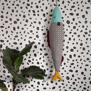 May include: A decorative fish-shaped hanging ornament. The fish has a teal head, a patterned body with red accents, and a yellow tail. It hangs from a chain against a white background with black polka dots. A green plant is in the lower left corner.