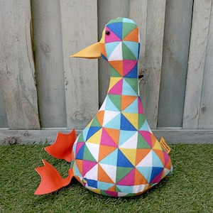 May include: A vibrant, geometric patterned duck soft toy. The duck has a patchwork design of triangles in various colours, including blue, green, yellow, orange, and pink. It has an orange beak and feet, and is sitting on green grass.