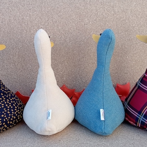 May include: Four plush goose-shaped doorstops in different patterns. The doorstops are lined up in a row from left to right: black with gold polka dots, white with a subtle texture, blue, and red plaid.