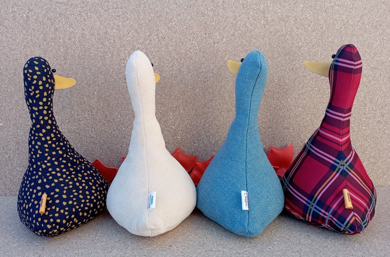 May include: Four plush goose-shaped doorstops in different patterns. The doorstops are lined up in a row from left to right: black with gold polka dots, white with a subtle texture, blue, and red plaid.
