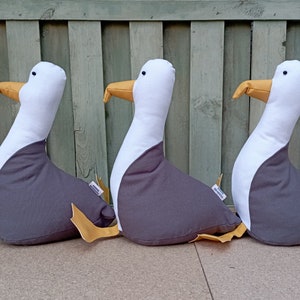 Può includere: Tre cuscini di peluche a forma di gabbiano grigi e bianchi con becco e piedi gialli. I cuscini sono disposti in fila su una superficie marrone chiaro.