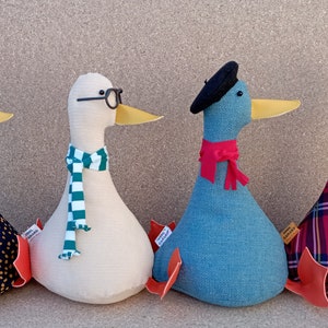 May include: Four plush toy ducks in different colors and patterns. One duck is wearing a party hat, another has a scarf and glasses, one has a beret and scarf, and the last one has a crown and plaid fabric.