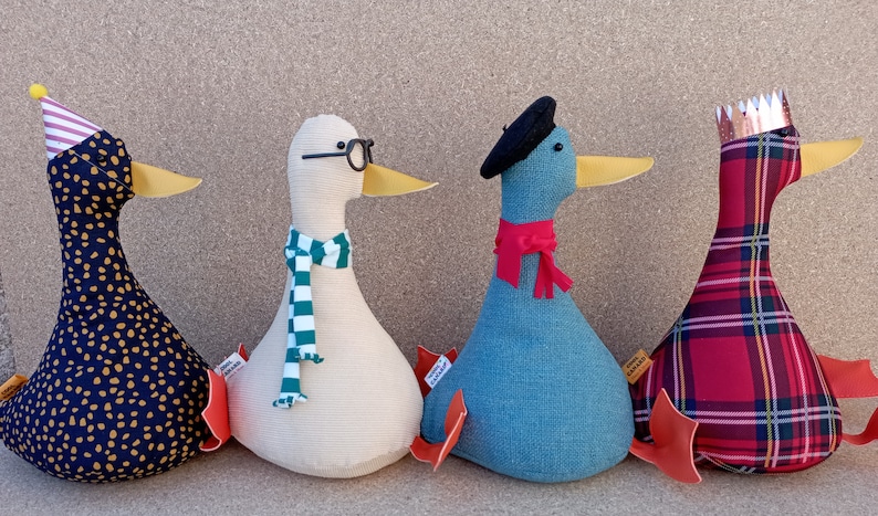 May include: Four plush toy ducks in different colors and patterns. One duck is wearing a party hat, another has a scarf and glasses, one has a beret and scarf, and the last one has a crown and plaid fabric.