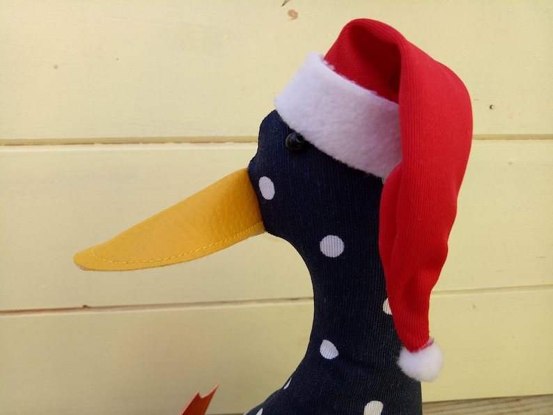 May include: A black and white polka dot duck wearing a red Santa hat with a white trim. The duck has a yellow beak and black eyes.