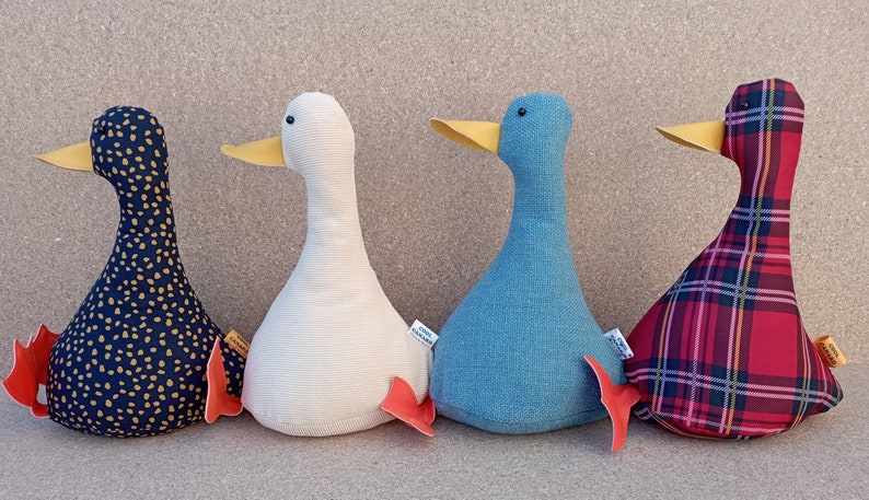 May include: Four plush toy ducks in different patterns. The ducks are sitting in a row on a brown surface. The first duck is black with gold polka dots, the second is white, the third is blue, and the fourth is red with a plaid pattern.