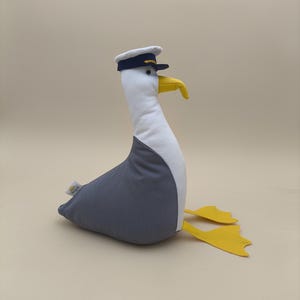 Könnte beinhalten: Ein Stofftier in Form einer Möwe mit weißem Kopf und Hals, grauem Körper und gelben Füßen und Schnabel. Die Möwe trägt eine marineblaue und weiße Kapitänsmütze. Das Spielzeug ist so konzipiert, dass es aufrecht sitzt.
