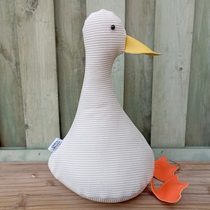 Puede incluir: Un juguete de peluche de ganso blanco y gris a rayas con un pico amarillo y patas naranjas. El ganso tiene un ojo negro y una pequeña etiqueta que dice "Cool + Chic".