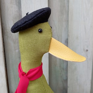 May include: A green fabric duck wearing a black beret and a red scarf. The duck has a yellow leather beak.
