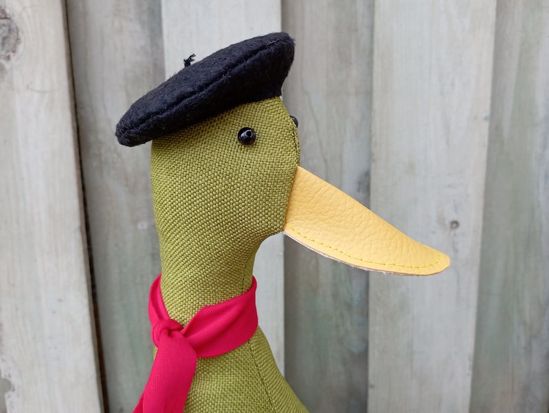 May include: A green fabric duck wearing a black beret and a red scarf. The duck has a yellow leather beak.